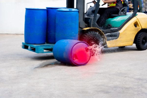 A forklift transporting a pallet of barrels with one barrel falling off and spilling on the ground