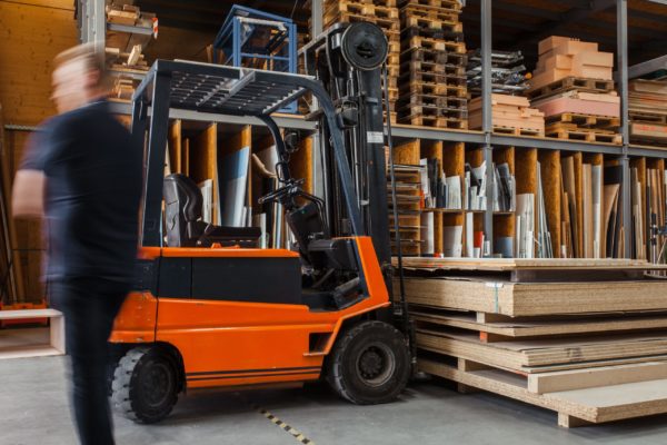 forklift-under-wood-sheets An orange forklift inside a warehouse with a worker walking past it