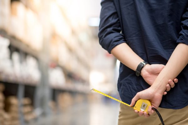 A person standing in a warehouse aisle and holding a tape measure behind their back