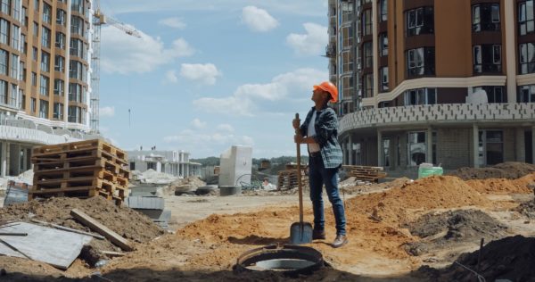 A worker on a construction site holding a shovel and wearing a hard hat while looking up to the sky