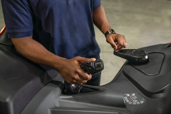 A stand-up forklift operator with one hand on the control joystick and the other on the steering knob