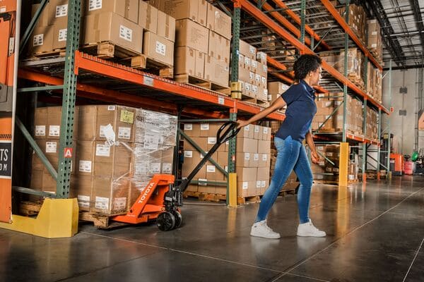 A warehouse worker pulling a pallet jack with a pallet from racking