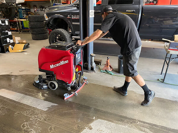 A worker scrubbing an automotive shop floor with a Factory Cat MicroMini walk-behind floor scrubber