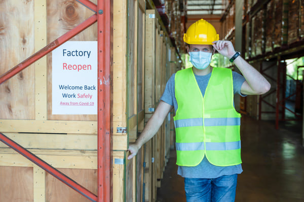 A warehouse worker wearing a hard hat, safety vest, and face mask