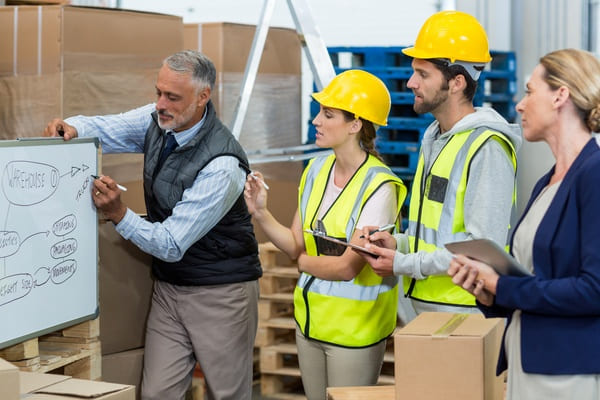warehouse-workers-having-a-meeting A supervisor writing on a whiteboard before a group of warehouse workers