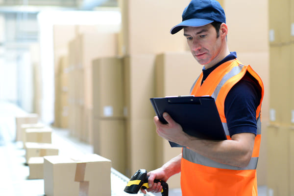 A warehouse worker in an orange safety vest scanning a box with an RF scanner while looking at a clipboard