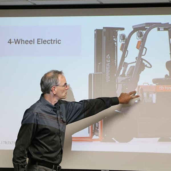 A forklift trainer pointing at a forklift on a projector screen in a classroom
