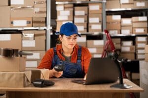 A warehouse worker scanning a box with an RF scanner while looking at a laptop