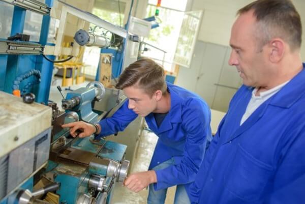 An older worker assisting a younger worker on a lathe machine