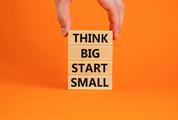 A hand stacking blocks that spell out "Think Big Start Small"