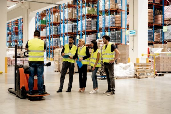 A forklift operator trainer stands on an electric pallet jack while giving instructions to operator trainees in a warehouse